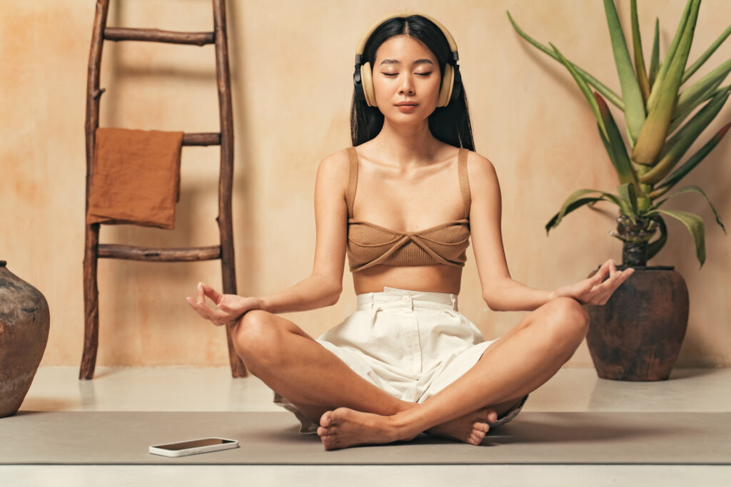 Asian woman in headphones meditating, sitting in yoga lotus pose, using meditation app on phone, keeping hands in mudra sign, listening to mantra in headphones with closed eyes, reaching zen
